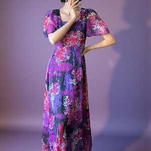 May include: A long, flowing dress with a vibrant floral pattern in shades of purple, pink, and black. The dress features short, flutter sleeves and a square neckline. The fabric appears to be lightweight and sheer, creating an elegant silhouette.