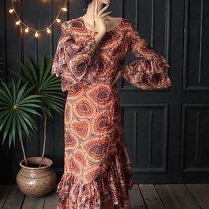 May include: A long, flowing dress with a vibrant, repeating pattern in shades of orange, brown, and black. The dress features long sleeves with flared cuffs and a ruffled hem. The dress is displayed on a mannequin.