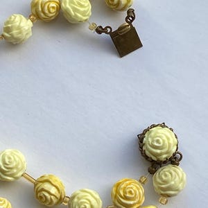 May include: A vintage necklace featuring cream and yellow rose-shaped beads. The beads are strung together with a delicate, transparent cord. The necklace has a bronze-colored clasp.