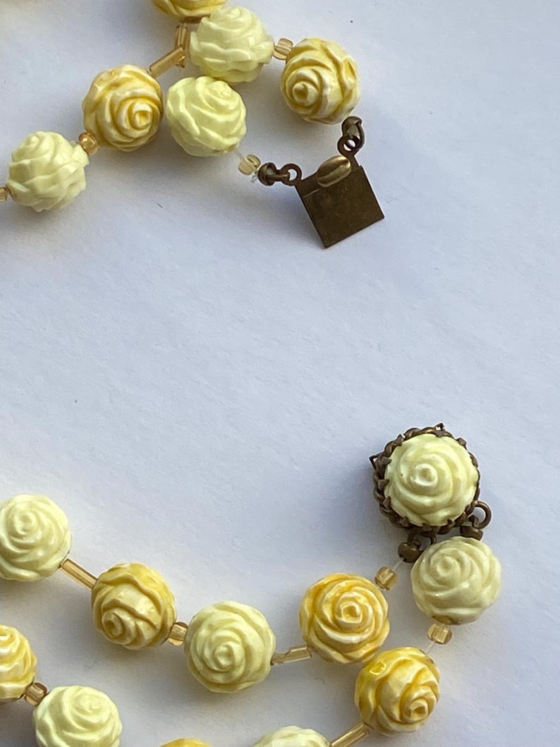 May include: A vintage necklace featuring cream and yellow rose-shaped beads. The beads are strung together with a delicate, transparent cord. The necklace has a bronze-colored clasp.