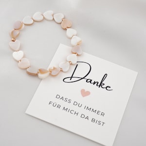Könnte beinhalten: Ein weißes und rosafarbenes Armband aus Herzperlen mit einem kleinen goldenen Herz-Anhänger. Das Armband liegt auf einer weißen Karte mit dem Text "Danke dass du immer für mich da bist" in Schwarz.
