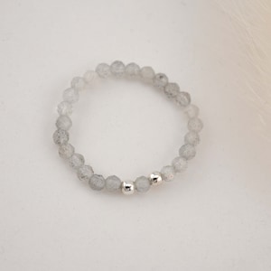 Könnte beinhalten: Ein graues Perlenarmband mit zwei silbernen Perlen. Das Armband besteht aus kleinen, runden Perlen mit einem gesprenkelten, wolkigen Aussehen.