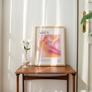 May include: A framed poster featuring an abstract design in shades of pink and yellow. The poster displays the text "ARIES" in black lettering and the dates "March 21 - April 19" in smaller black lettering. The poster is hanging on a wall above a wooden table.