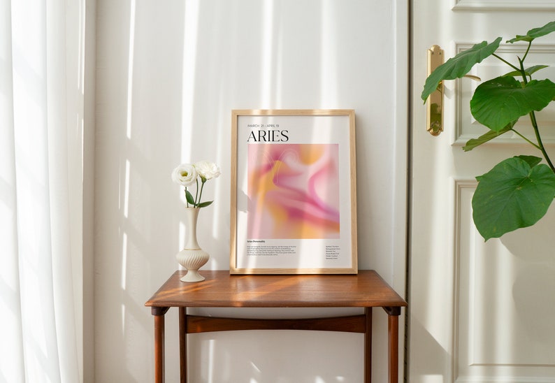 May include: A framed poster featuring an abstract design in shades of pink and yellow. The poster displays the text "ARIES" in black lettering and the dates "March 21 - April 19" in smaller black lettering. The poster is hanging on a wall above a wooden table.