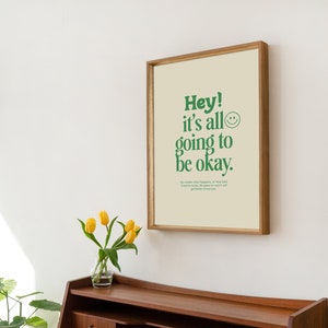 May include: A framed print with a light beige background and green text that reads "Hey! It's all going to be okay." A green smiley face is next to the text. The print is in a light wood frame and is hanging on a white wall.