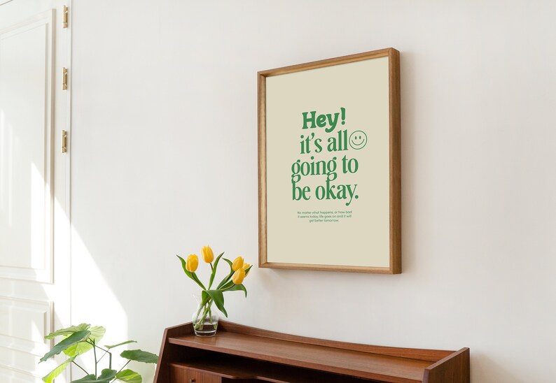 May include: A framed print with a light beige background and green text that reads "Hey! It's all going to be okay." A green smiley face is next to the text. The print is in a light wood frame and is hanging on a white wall.