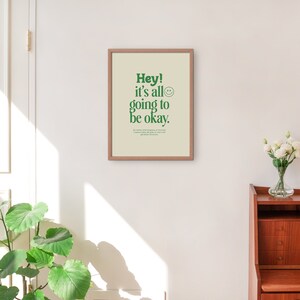 May include: A framed print with a white background and green text that reads "Hey! It's all going to be okay."  A smiley face icon is next to the text. The print is hanging on a white wall.