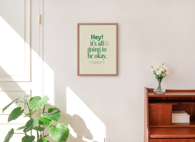 May include: A framed print with a white background and green text that reads "Hey! It's all going to be okay."  A smiley face icon is next to the text. The print is hanging on a white wall.