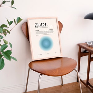 May include: A framed print with the word "aura" in black text on a white background. The print features a blue and green gradient circle in the center. The print is sitting on a wooden chair in a room with a white wall and a wooden table with a lamp.