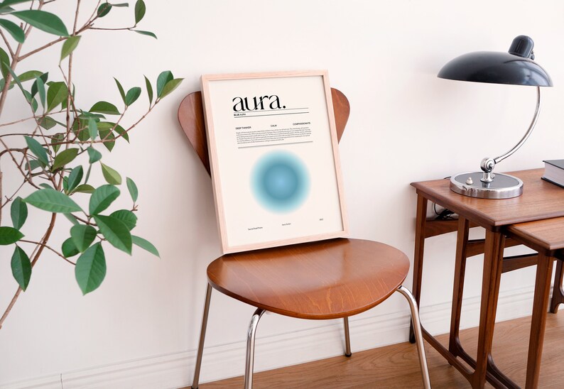 May include: A framed print with the word "aura" in black text on a white background. The print features a blue and green gradient circle in the center. The print is sitting on a wooden chair in a room with a white wall and a wooden table with a lamp.