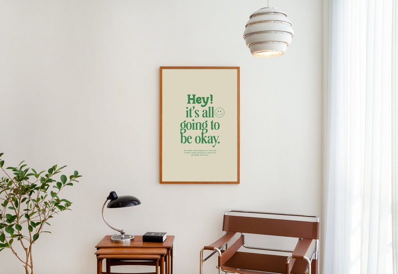 May include: A framed print with a white background and green text that reads "Hey! It's all going to be okay."  A smiley face is included in the text. The print is hanging on a white wall.