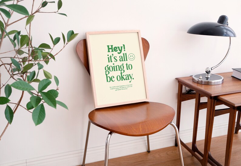 May include: A framed print with a light beige background and green text that reads "Hey! It's all going to be okay." A smiley face is next to the text. The print is sitting on a wooden chair.