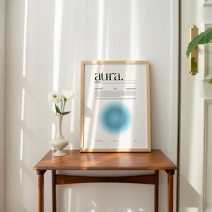 May include: A framed print with the word "aura" in black text on a white background. The print features a blue circle with a gradient effect. The print is hanging on a white wall above a wooden table with a white vase and white flowers.