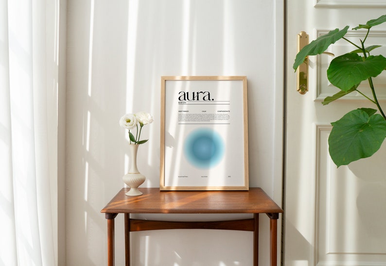 May include: A framed print with the word "aura" in black text on a white background. The print features a blue circle with a gradient effect. The print is hanging on a white wall above a wooden table with a white vase and white flowers.