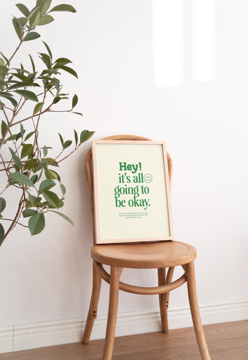 May include: A framed print with a light green background and a green message that reads "Hey! It's all going to be okay." The print is sitting on a wooden chair in front of a white wall.