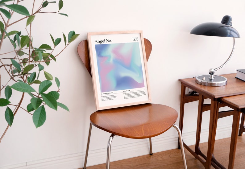 May include: A framed print with a light blue, pink, and purple abstract design. The print has the text "Angel No. 555" at the top. The print is sitting on a wooden chair in front of a white wall.