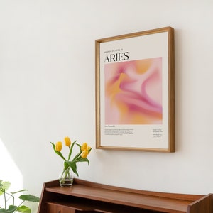 May include: A framed poster featuring a pink and yellow abstract design. The poster displays the text "MARCH 21 - APRIL 19" and "ARIES" in black lettering. The poster is hanging on a white wall.