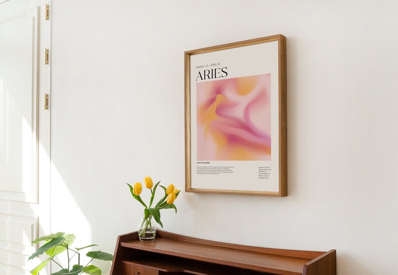 May include: A framed poster featuring a pink and yellow abstract design. The poster displays the text "MARCH 21 - APRIL 19" and "ARIES" in black lettering. The poster is hanging on a white wall.