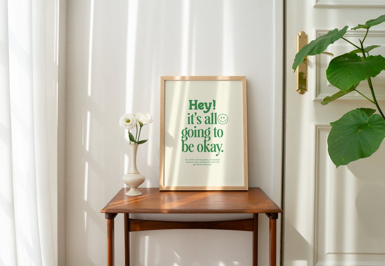 May include: A framed print with a white background and green text that reads "Hey! It's all going to be okay."  A smiley face is next to the text. The print is on a wooden table with a vase of white flowers.