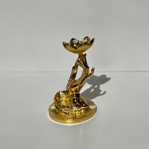 May include: A gold metal ring holder shaped like a hand holding a lotus flower. The ring holder is on a white surface.