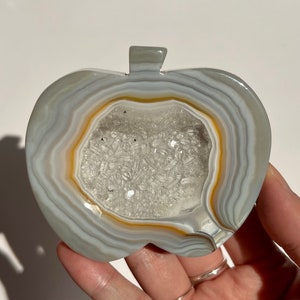 May include: A white agate geode carved into the shape of an apple. The geode has a natural opening that reveals a cluster of clear quartz crystals.