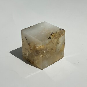 Puede incluir: Un objeto decorativo translúcido en forma de cubo con un patrón veteado blanco y beige. El cubo está hecho de un material sólido y tiene una superficie lisa y pulida. El objeto proyecta una sombra sobre una superficie blanca.