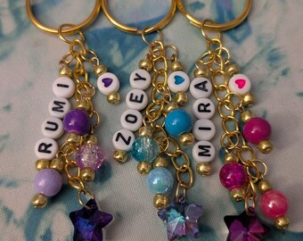 May include: Three personalized keychains with gold rings. Each keychain features a name spelled out with white letter beads, accented with colorful beads and a star-shaped charm. The names are Rumi, Zoey, and Mira.