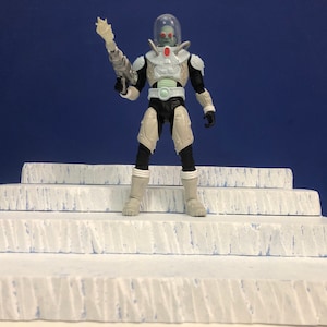 May include: A white and black action figure of Mr. Freeze, a supervillain from Batman comics, standing on a set of white steps. He is wearing a white and black suit with a clear helmet and a freeze ray in his right hand.