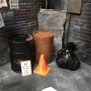 Könnte beinhalten: Ein Diorama einer urbanen Gasse mit Backsteinmauern, Metallblechen und einem Betonboden. Es gibt zwei schwarze Fässer, ein rostbraunes Fass, einen orangefarbenen Verkehrskegel, einen schwarzen Müllsack und ein Schild mit der Aufschrift "NO VEHICLES BEYOND THIS POINT."