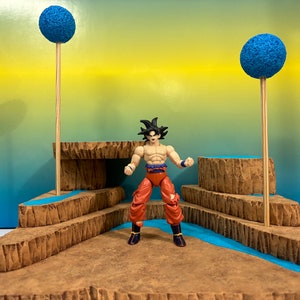 May include: An action figure of a character with black hair, wearing a red and blue outfit, stands on a brown platform with two blue spheres on wooden poles. The platform has a blue and brown layered design.