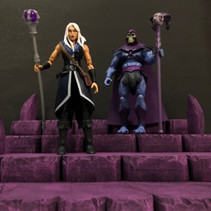 May include: Two action figures, a female figure with white hair and a blue and black outfit holding a staff with a purple orb, and a male figure with purple skin and a purple cape holding a staff with a skull, standing on a purple rock formation.