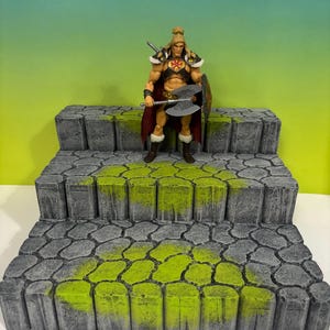 May include: A blonde action figure with a red cape, brown boots, and a chest emblem, holding a silver double-bladed axe and a shield. The figure stands on a three-tiered gray stone staircase display base with textured moss-green painted details.