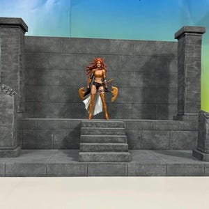 Puede incluir: Un escenario de diorama de piedra gris con una figura de acción femenina. El escenario tiene una plataforma central con escalones, flanqueada por pilares y elementos decorativos. La figura lleva una armadura dorada y sostiene dos hachas.