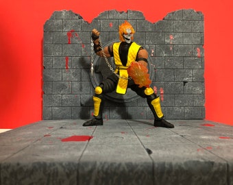 Op de afbeelding: Een geel en zwart action figure van een personage met een schedelgezicht en vlammen op zijn hoofd. De figuur houdt een ketting vast met een punt aan het einde. De figuur staat voor een grijze bakstenen muur met rode bloedspatten.