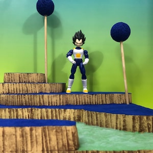 May include: A blue and yellow action figure of a character from the anime Dragon Ball Z standing on a multi-level platform with blue and brown accents. The platform is surrounded by green grass and two blue trees.