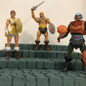 Op de afbeelding: Drie action figuren uit de Masters of the Universe franchise. De figuren zijn He-Man, She-Ra en Man-At-Arms. He-Man houdt een zwaard en een schild vast, She-Ra houdt een zwaard en een schild vast en Man-At-Arms houdt een knots vast. De figuren staan op een stenen platform.