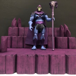 Op de afbeelding: Een paars en zwart action figure van Skeletor uit Masters of the Universe, staand op een paars rotsplatform. Skeletor houdt een staf vast met een schedel erop.