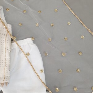 May include: White outfit with gold sequin embellishments. The outfit includes a top with gold sequins, a skirt, and a sheer white veil with gold floral accents and a gold trim. The outfit is likely for a special occasion.