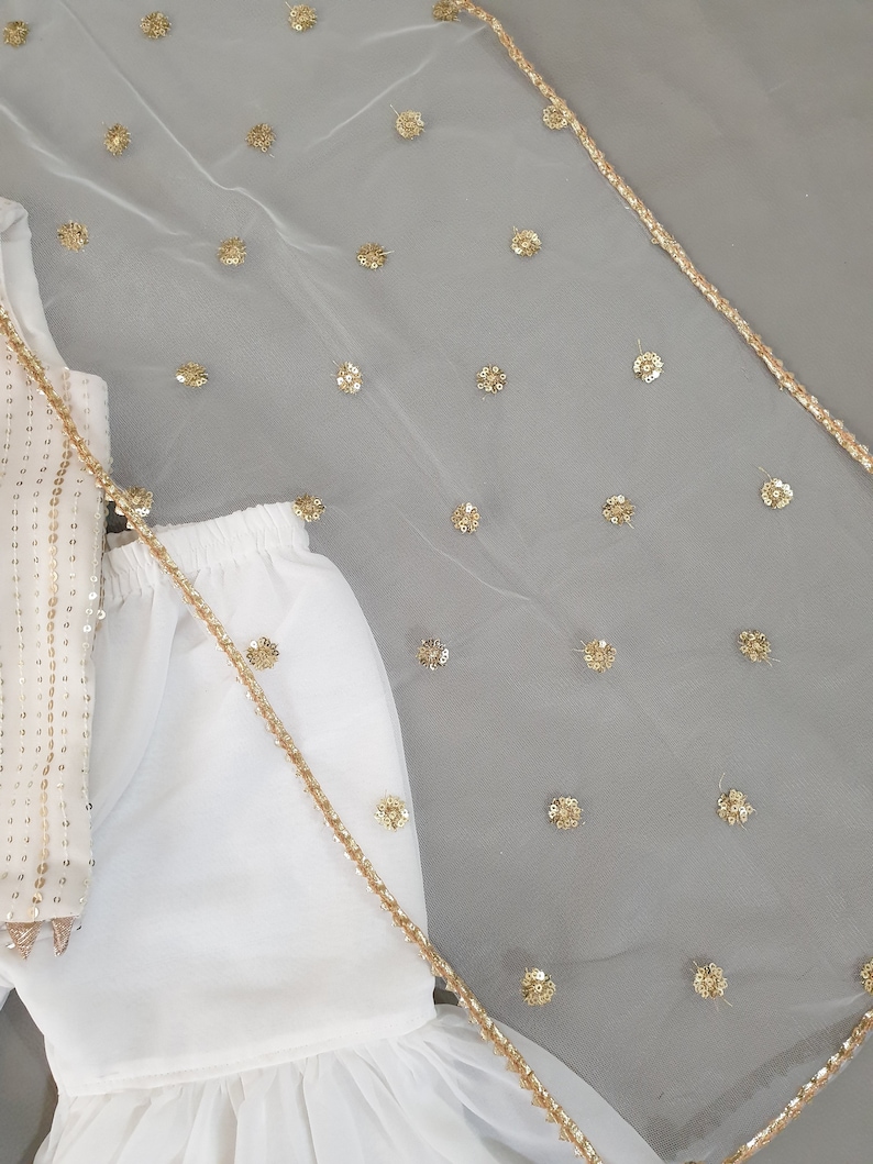 May include: White outfit with gold sequin embellishments. The outfit includes a top with gold sequins, a skirt, and a sheer white veil with gold floral accents and a gold trim. The outfit is likely for a special occasion.