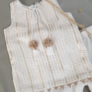 May include: White sleeveless top and pants set with gold sequin embellishments. The top features a keyhole neckline with tassel ties and gold pom-poms. The pants have a tiered design. A sheer white scarf with gold accents completes the outfit.