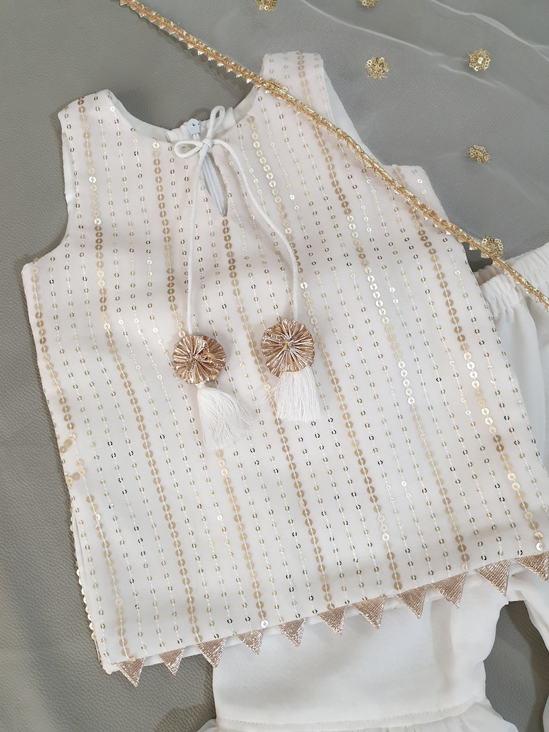 May include: White sleeveless top and pants set with gold sequin embellishments. The top features a keyhole neckline with tassel ties and gold pom-poms. The pants have a tiered design. A sheer white scarf with gold accents completes the outfit.
