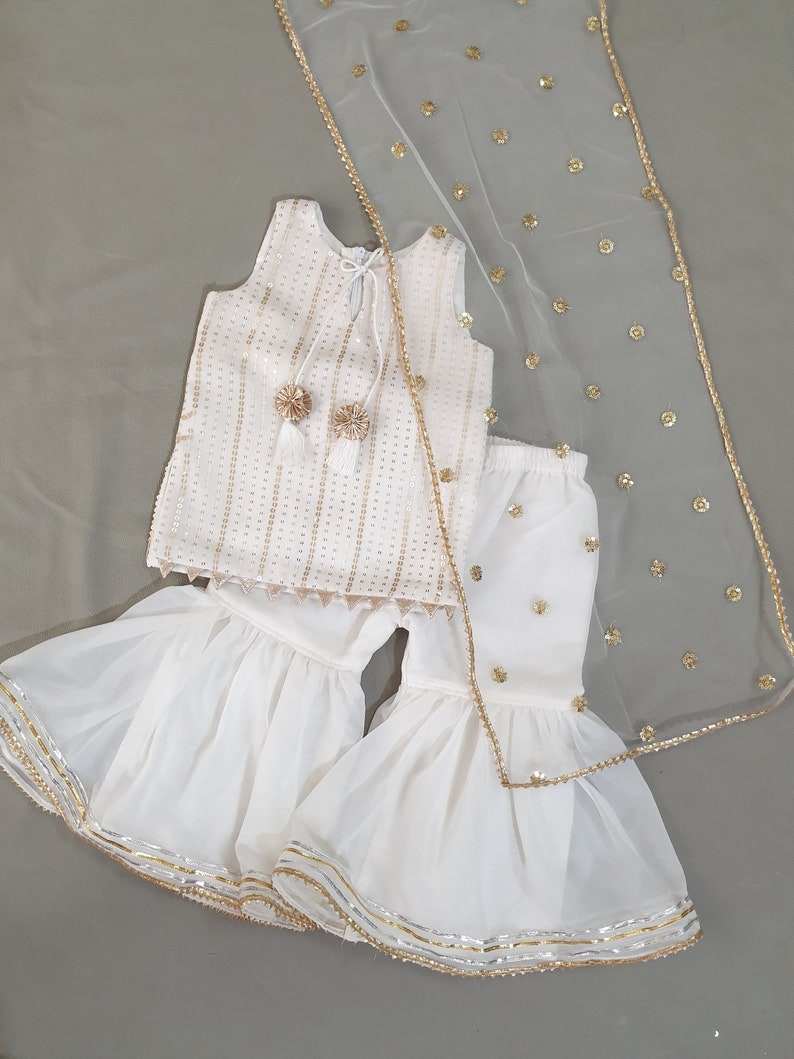 May include: A white, sleeveless outfit with gold accents, including a top, flared pants, and a sheer scarf. The top and pants are embellished with gold sequins. The scarf has gold embellishments and a gold trim. The outfit is likely for a special occasion.