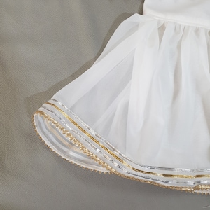 May include: White formal wear set with gold and silver trim. The top is embellished with sequins and triangular gold accents. The skirt has a layered, flowing design with gold and silver trim along the hemline. The outfit is set against a gray background.