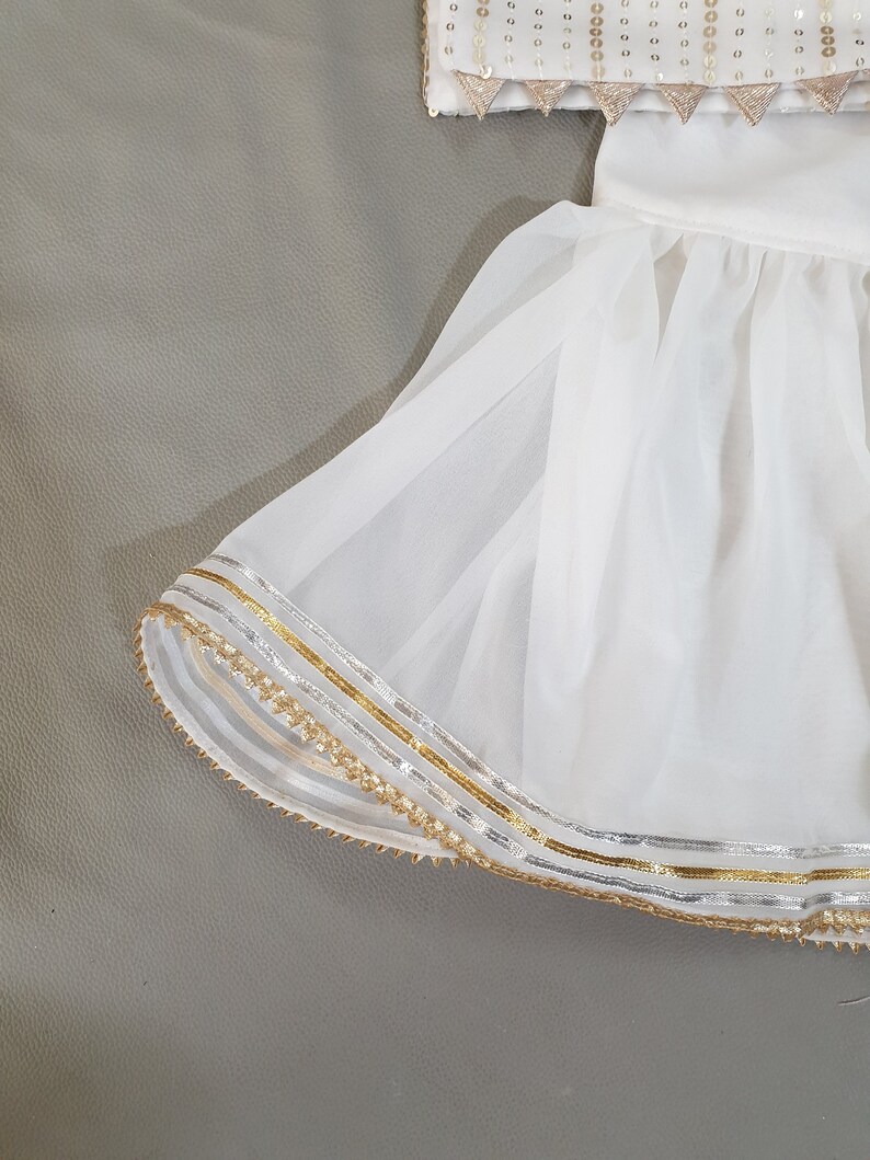 May include: White formal wear set with gold and silver trim. The top is embellished with sequins and triangular gold accents. The skirt has a layered, flowing design with gold and silver trim along the hemline. The outfit is set against a gray background.