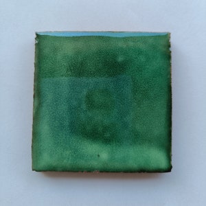 Puede incluir: Un azulejo cuadrado con un esmalte verde. El esmalte tiene una textura moteada y un ligero tono azul.
