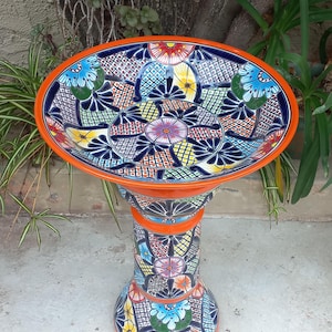 May include: A ceramic birdbath featuring a vibrant, hand-painted Talavera-style pattern with intricate geometric shapes and floral motifs in shades of blue, yellow, red, and green. The basin and pedestal have a bright orange rim and base.