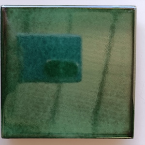 May include: Square, glossy, green ceramic tile with a darker green rectangular design in the center. The tile has a reflective surface and a smooth, polished finish. The edges are clean and well-defined, suggesting a high-quality craft.