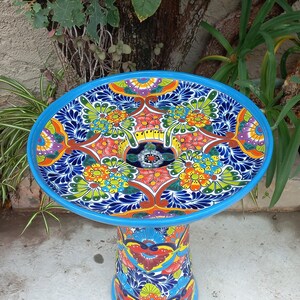 May include: A round, pedestal-style birdbath featuring a vibrant, hand-painted Talavera-inspired pattern. The surface displays intricate floral motifs in shades of blue, orange, yellow, green, and red, finished with a solid light blue rim.