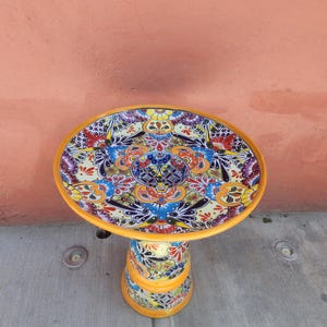 May include: A round birdbath featuring a vibrant, multi-colored floral and geometric pattern in shades of yellow, blue, red, and black. The basin and the cylindrical pedestal are decorated with intricate hand-painted designs and a solid yellow rim.