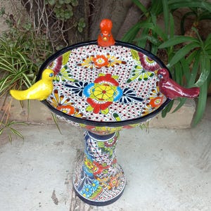 May include: A ceramic birdbath featuring a hand-painted Talavera-style floral pattern in vibrant red, yellow, blue, and green. The basin has a black rim and three attached ceramic birds in solid yellow, orange, and maroon perched on the edge.